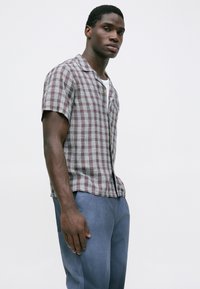 Uomo che indossa una camicia a maniche corte a quadri marroni e bianchi sopra una maglietta bianca, abbinata a pantaloni blu, in piedi davanti a uno sfondo bianco semplice.