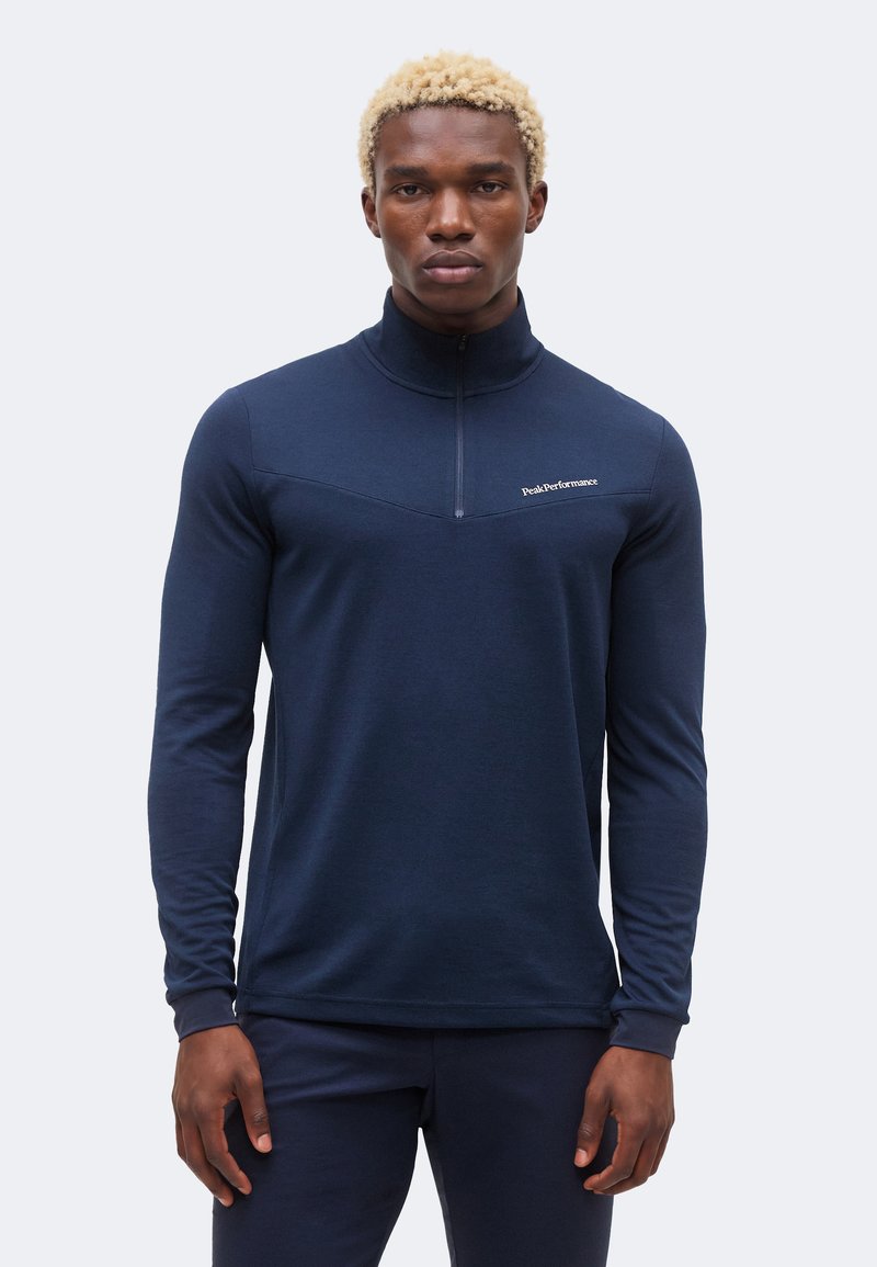 Άνδρας με κοντά σγουρά ξανθά μαλλιά φοράει μπλε σκουρόχρωμο μακρυμάνικο τοπ με φερμουάρ έως τη μέση και αντίστοιχο παντελόνι, στέκεται μπροστά σε απλό φωτεινό φόντο.