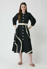Knopfhemd, schwarzes Kleid mit weißen Akzentlinien, kurzen Ärmeln und Taillengurt. Verfügt über Taschen und einen fließenden Rock. Getragen mit braunen Sandalen.