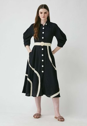 Knopfhemd, schwarzes Kleid mit weißen Akzentlinien, kurzen Ärmeln und Taillengurt. Verfügt über Taschen und einen fließenden Rock. Getragen mit braunen Sandalen.