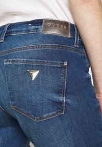 Modré džíny z denimu s přízdobou stříbrného loga "GUESS", trojúhelníkovým logem na zadní kapse a kontrastním prošíváním.