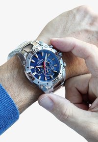 Montre en acier inoxydable argenté avec un cadran bleu, fonctionnalités de chronographe, aiguilles lumineuses et un bracelet texturé, en cours d'ajustement sur un poignet.