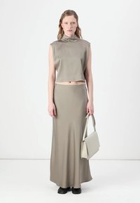 Ensemble en deux pièces de couleur olive atténuée ; top sans manches, court avec un col haut, et une longue jupe ajustée. Le mannequin tient un sac à main assorti.