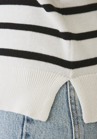 Maglia a righe bianco e nero in maglia, con una texture a costine. Presenta un orlo spaccato, abbinata a jeans in denim azzurro chiaro. Design semplice e funzionale.