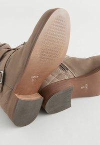Botas de tobillo marrones con un empeine de cuero liso, detalles acolchados y un tacón bajo tipo bloque. Suela con dibujo para mayor agarre. Branding visible en la suela.