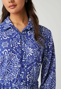 Blaues Hemd mit Knopfverschluss, das ein weißes florales und abstraktes Muster zeigt. Glattes Gewebe mit Kragen und langen Ärmeln. Die Knöpfe passen farblich zum Stoff.