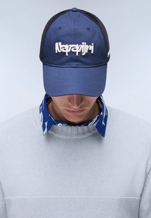 Hombre con gorra azul marino con el logo blanco de "Napapijri", camisa azul con estampado y cuello debajo de un jersey de punto gris claro, cabeza inclinada hacia abajo.