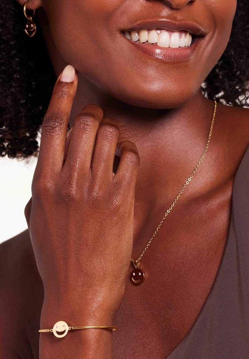 Gouden ketting met een bruine smiley-hanger, bijpassende gouden armband met een smiley-charme, tegen een zachte huidtint.