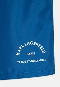Mėlynas audinys su lygiu paviršiumi, ant kurio yra balta užrašyta tekstas "KARL LAGERFELD PARIS 21 RUE ST-GUILLAUME" išlenktame dizaine.