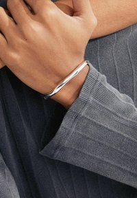 Bracelet en métal argenté avec un design lisse et torsadé, porté sur une main avec une manche de pull à côtes gris foncé et texturé.