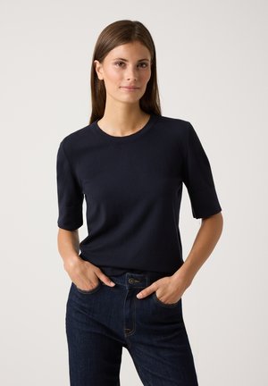 Marineblaues kurzärmliges Oberteil aus Baumwolle, runder Halsauschnitt, taillierte Form. Getragen mit dunklen Jeans.