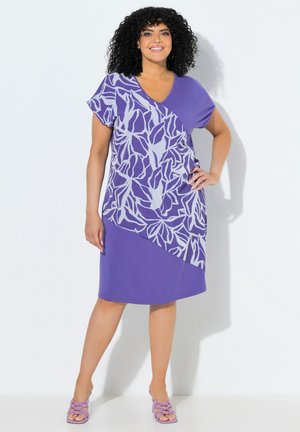 Frau mit lockigem schwarzen Haar, die ein violettes knielanges Kleid mit leichtem Blumenmuster und violette offene Zehenschuhe trägt, lächelnd mit der Hand in der Hüfte.