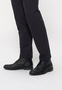 Czarne skórzane botki za kostkę o gładkim wykończeniu, okrągłym nosku i sznurowanym designie. W połączeniu z wąskimi ciemnymi spodniami.