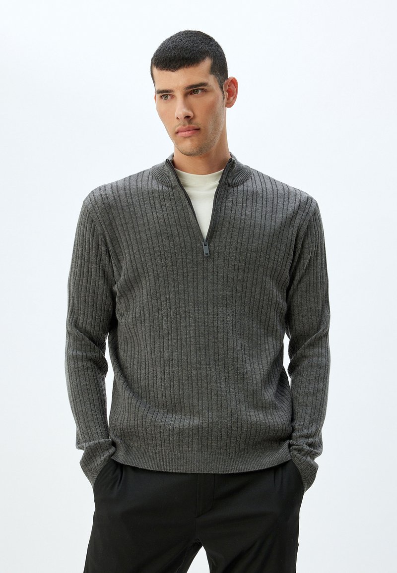 Suéter gris acanalado con un cuello medio con cremallera, mangas largas y un diseño ajustado. La tela tiene una apariencia texturizada y un acabado suave.