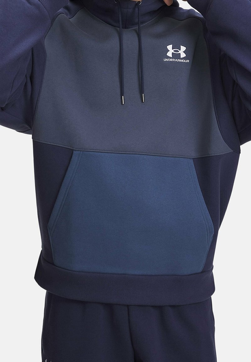Personne portant un sweat à capuche Under Armour bicolore bleu avec poche avant et cordons pour ajuster la capuche.
