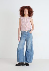 Haut sans manches rose avec un motif texturé ; jean bleu taille haute à jambes larges ; bottines noires. Fond épuré avec des tons neutres.