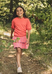 Koraalroze top met textuur en pofmouwen, gecombineerd met een gelaagde roze bloemenrok. Neutrale sneakers maken de outfit af. Natuurlijke buitenomgeving.