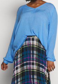 Vrouw in een losse blauwe lange mouwtop, ingestopt in een hooggeplooide geruite rok met groen, roze, blauw en zwarte strepen.