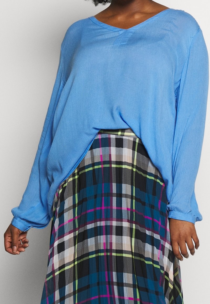 Vrouw in een losse blauwe lange mouwtop, ingestopt in een hooggeplooide geruite rok met groen, roze, blauw en zwarte strepen.