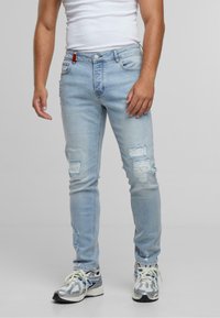 Homme portant un jean skinny déchiré bleu clair et des baskets blanches, se tenant contre un fond gris uni.