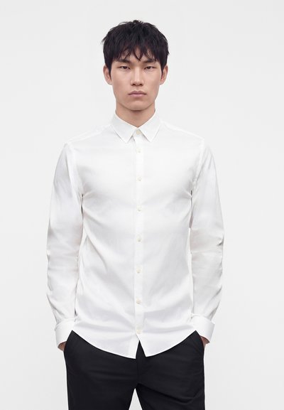 Man wearing a fitted white button-up shirt with a collar and black pants, standing with hands in pockets against a plain background.