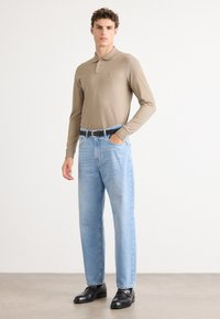 Beige hosszú ujjú pólóing kis logóval, világos kék egyenes szárú farmernadrággal és fekete bőrcipővel, semleges háttér előtt állva.