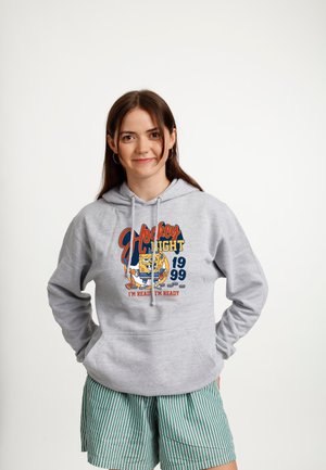 Ung kvinde iført en grå hættetrøje med SpongeBob hockey-grafik og grønne stribede shorts, stående med hænderne bag ryggen.