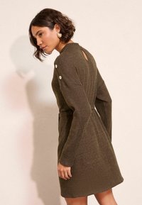 Abito in maglia verde oliva con maniche lunghe, vita con cintura e dettaglio a fessura sul retro. Presenta dei bottoni decorativi sulle spalle. Texture morbida.