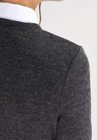 Cardigan grigio a maglia con una finitura testurizzata e una forma leggermente attillata. Presenta una camicia bianca colletto sotto, vista da dietro.