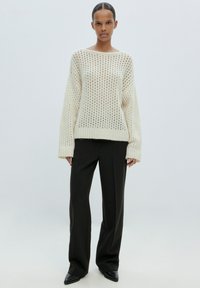 Grobstrickpullover in Creme mit offener Maschenstruktur, weiten Ärmeln und lockerer Passform, kombiniert mit schwarzen, weiten Hosen und spitzen Absätzen.