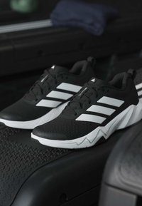 Μαύρα και λευκά παπούτσια τρεξίματος Adidas με τρεις λευκές ρίγες, τοποθετημένα σε διάδρομο με σκοτεινό φόντο.