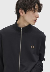 Felpa nera con zip e colletto alto, con logo in foglia d'oro sul lato sinistro del petto. Tessuto morbido, design aderente e polsini a costine.