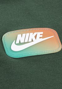 Patch du logo Nike sur un fond en tissu vert. Le patch présente un dégradé de couleurs, avec des lettres blanches et une finition brillante.