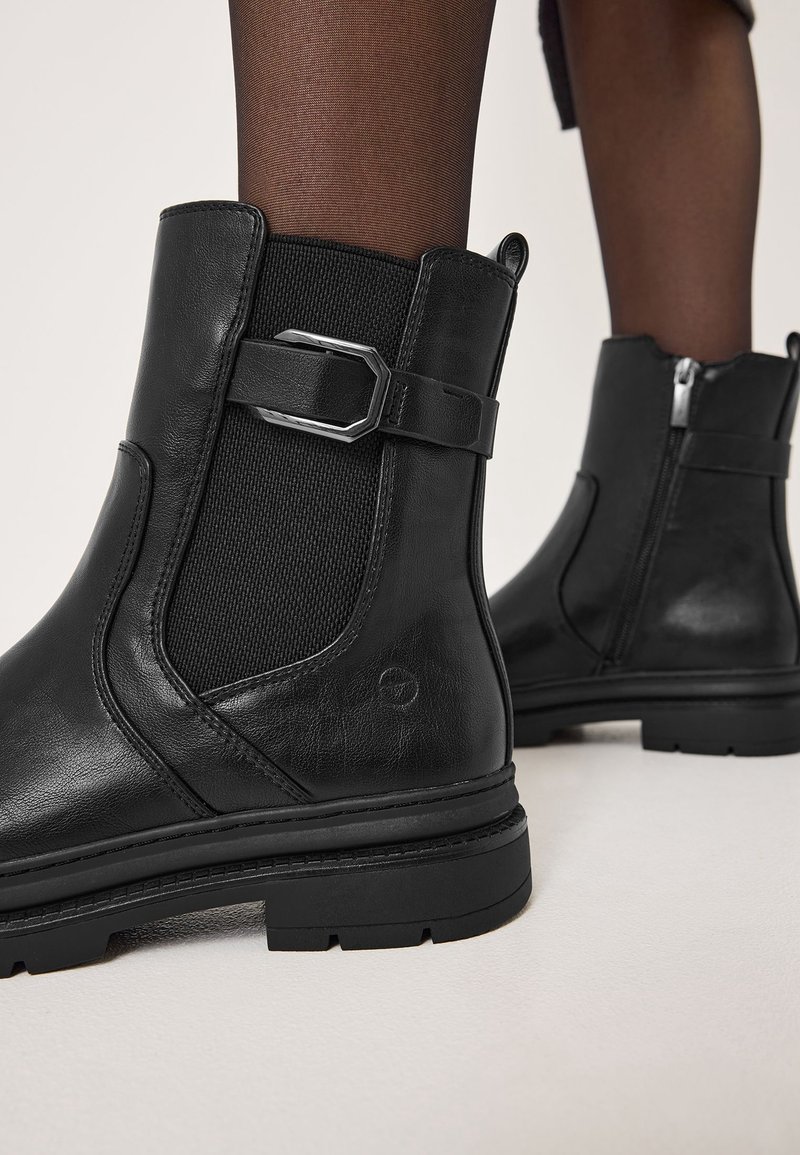 Bottines en cuir noir avec panneaux latéraux élastiques, boucle décorative et fermeture éclair latérale, portées avec des collants foncés transparents sur fond clair.