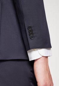 Navy suit jacket close-up with four black buttons on the cuff. White shirt cuff visible underneath, featuring curved hems. Smooth fabric texture.