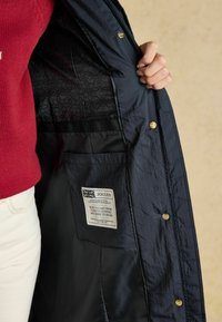 Námorná nepremokavá bunda s hladkým povrchom, ozdobená zlatými patentmi, prešívanou čiernou podšívkou a štítkom s informáciami o značke.