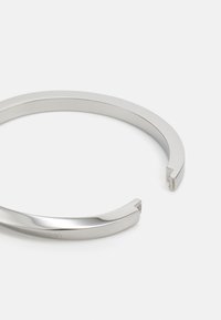 Bracciale cuff in metallo argento con un design sleek e aperto. Texture liscia, forma curvata con una leggera apertura, che mette in mostra semplicità ed eleganza.