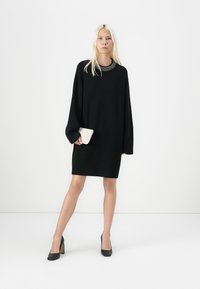 Svart oversized strikket kjole med ribbestrikket kant og mansjetter, med en perlepyntet utringning, parret med en liten hvit clutch og svarte hæler.