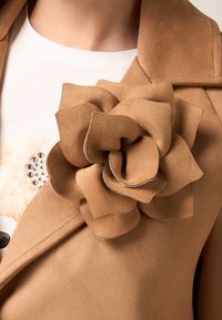 Abrigo de gamuza color beige con un gran aplique floral en la solapa izquierda. La flor presenta pétalos superpuestos y un ligero bordado, añadiendo textura.
