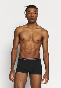 Sorte boxershorts med en hvid og rød "TOMMY HILFIGER" logo-linning, lavet af et glat, strækbart stof. Modellen står foran en ensfarvet baggrund.