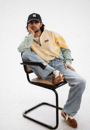 Ung mand iført briller, kasket og farveblok-strik sidder med korslagte ben på en moderne stol med flet-sæde og hviler sit ansigt på hånden.