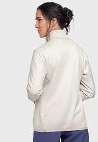 Helle beige Jacke mit hohem Kragen, langen Ärmeln, glatter Textur und taillierter Passform. Der Rücken hat ein dezentes Muster und wird mit blauen Hosen getragen.