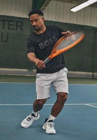 T-shirt de sport noir avec un grand logo "BOSS", short blanc et chaussures de tennis blanches avec des accents noirs et verts, maniant une raquette orange.