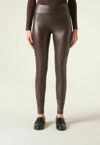 Hnědé legíny z umělé kůže s vysokým pasem, které mají hladkou a lesklou texturu a přiléhavý střih. Jsou nošeny černé slip-on boty.