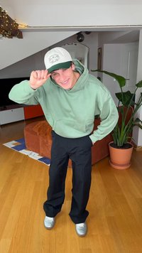 Groene hoodie, zwarte broek en zilveren sneakers. Draagt een witte pet met groene accenten. Binnenomgeving met zichtbare planten en meubels.