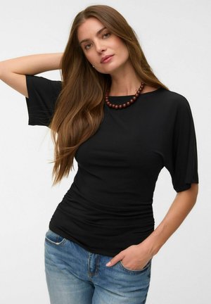 Femme aux longs cheveux bruns portant un haut noir à manches courtes, un jean bleu et un collier de perles, posant avec une main derrière la tête.