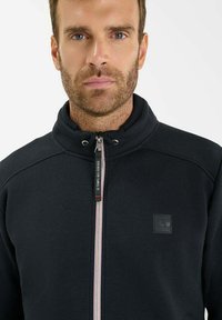 Uomo con capelli corti castani e barba di tre giorni, che indossa una giacca nera con zip e colletto alto, su uno sfondo bianco uniforme.