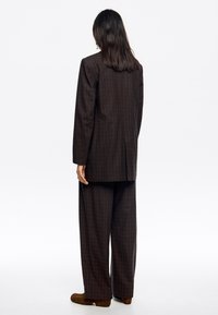 Completo a quadri marrone con giacca strutturata e pantaloni a gamba larga. Presenta un motivo a griglia blu, tessuto strutturato e una fessura centrale sul retro della giacca.
