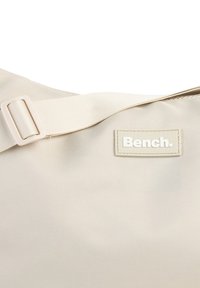 Beige Stoff-Umhängetasche mit glatter Textur, einem verstellbaren Tragegurt und einem kleinen rechteckigen Etikett mit der Aufschrift "Bench." in Weiß.