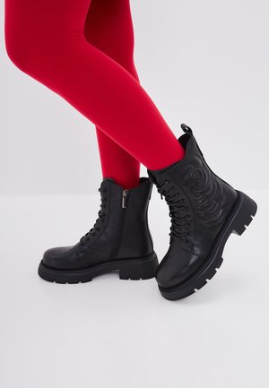 Schwarze Schnürstiefel im Kampfboots-Stil mit seitlichen Reißverschlüssen und geprägt-blumigem Design, getragen zu leuchtend roten Strumpfhosen vor einem schlichten hellen Hintergrund.